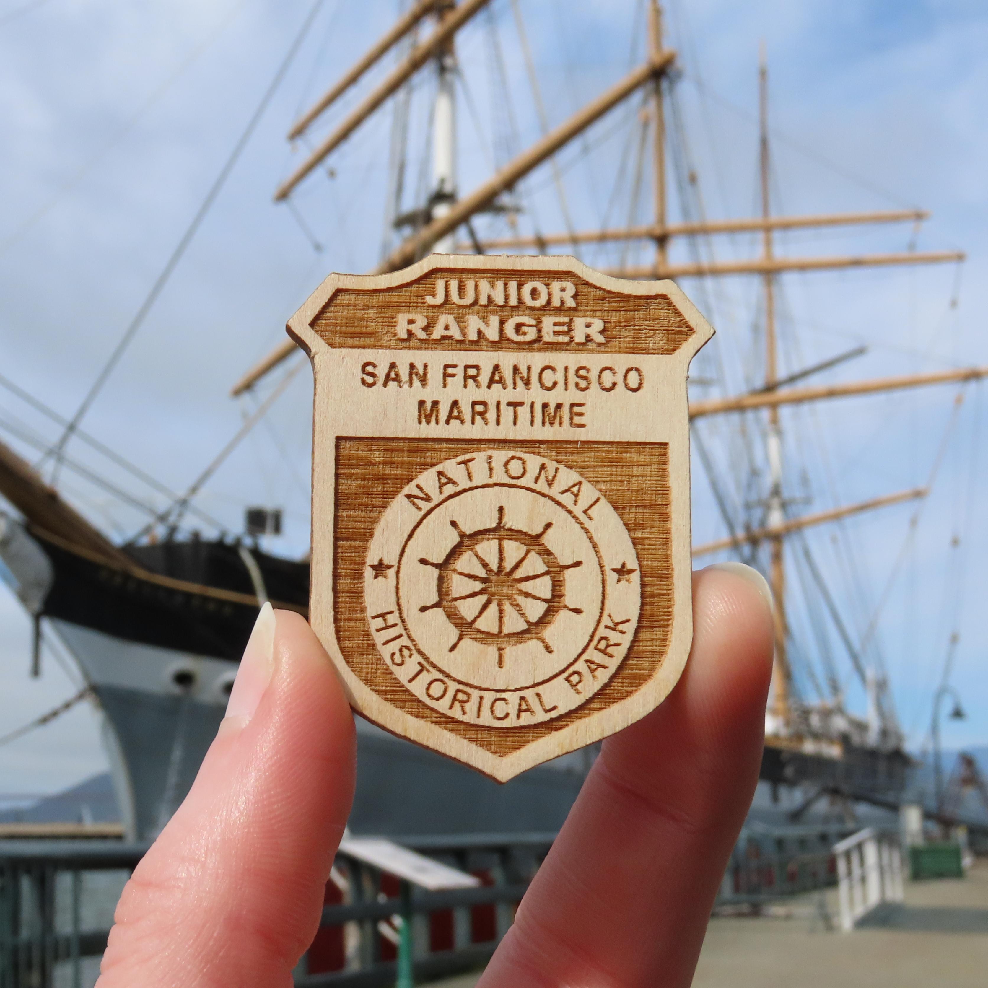 a San Francisco Maritime Junior Ranger Badge is held up, with a ship visible in the background.