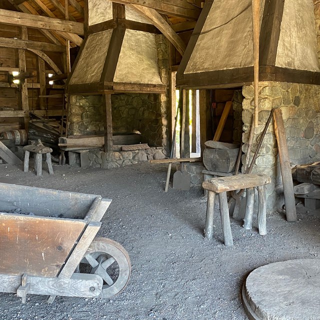 Building interior with two chimneys, gravel floor, stool, wheelbarrow, and other blacksmith tools