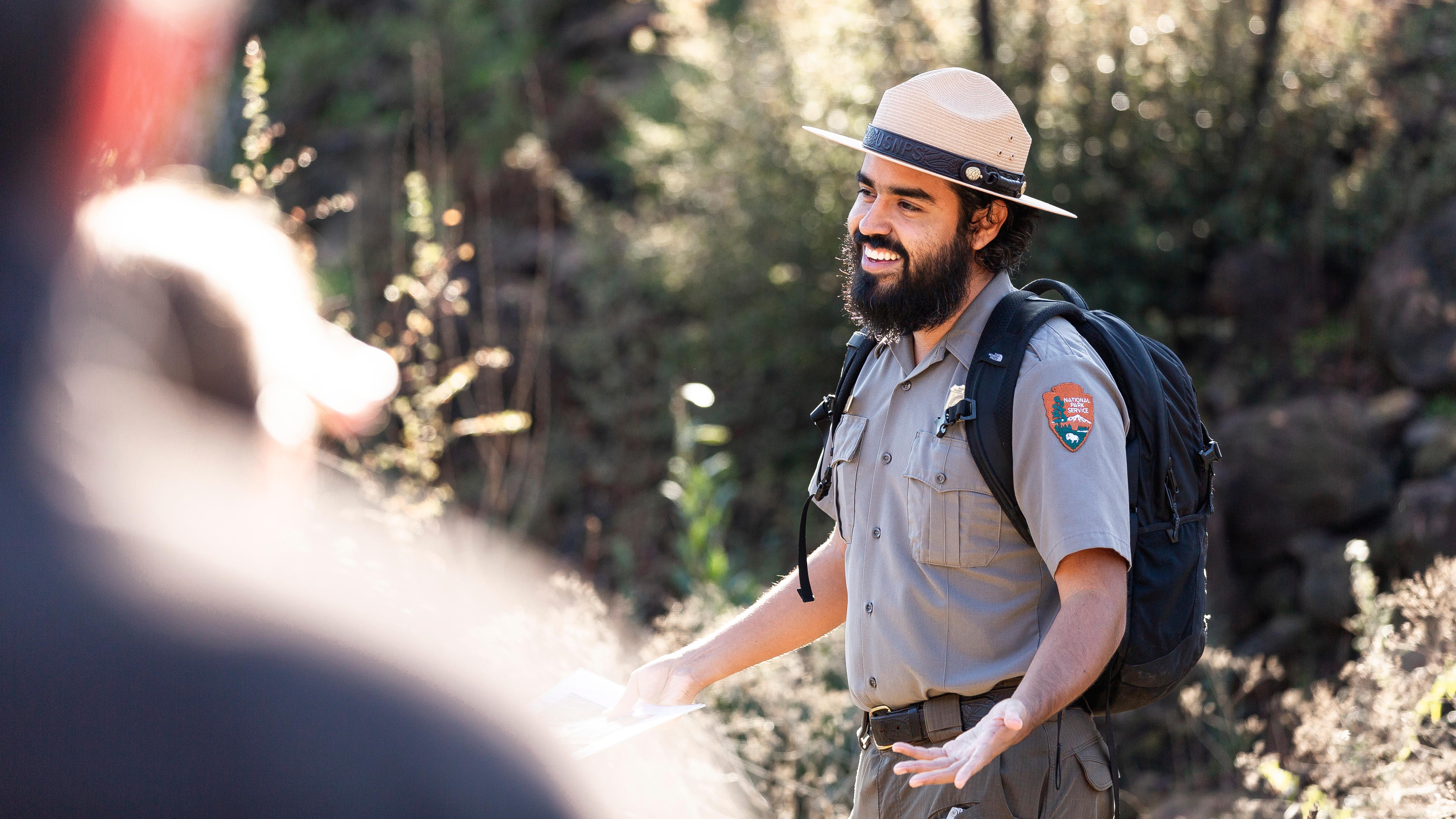 Ranger with backpack smiling with his arms open in the middle of talking.