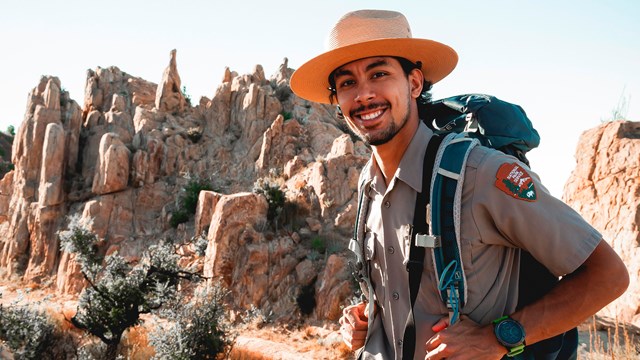 A smiling ranger standing with a backpack on in front of rocky outcrop.