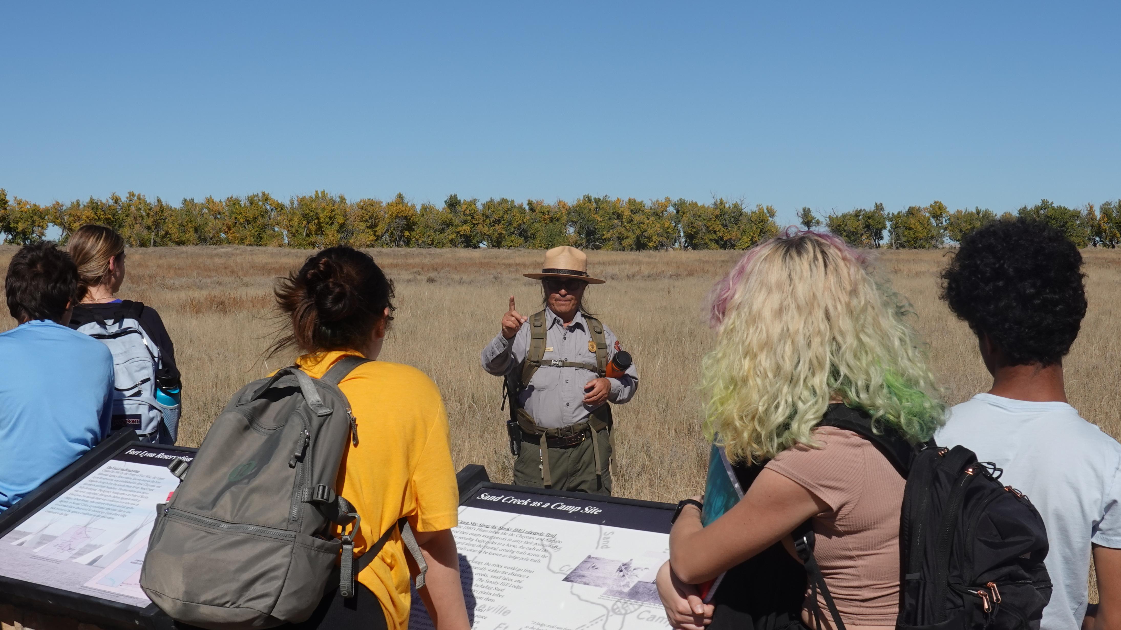 A park ranger talks to people in front of an open plain. A line of trees is in the distance. 