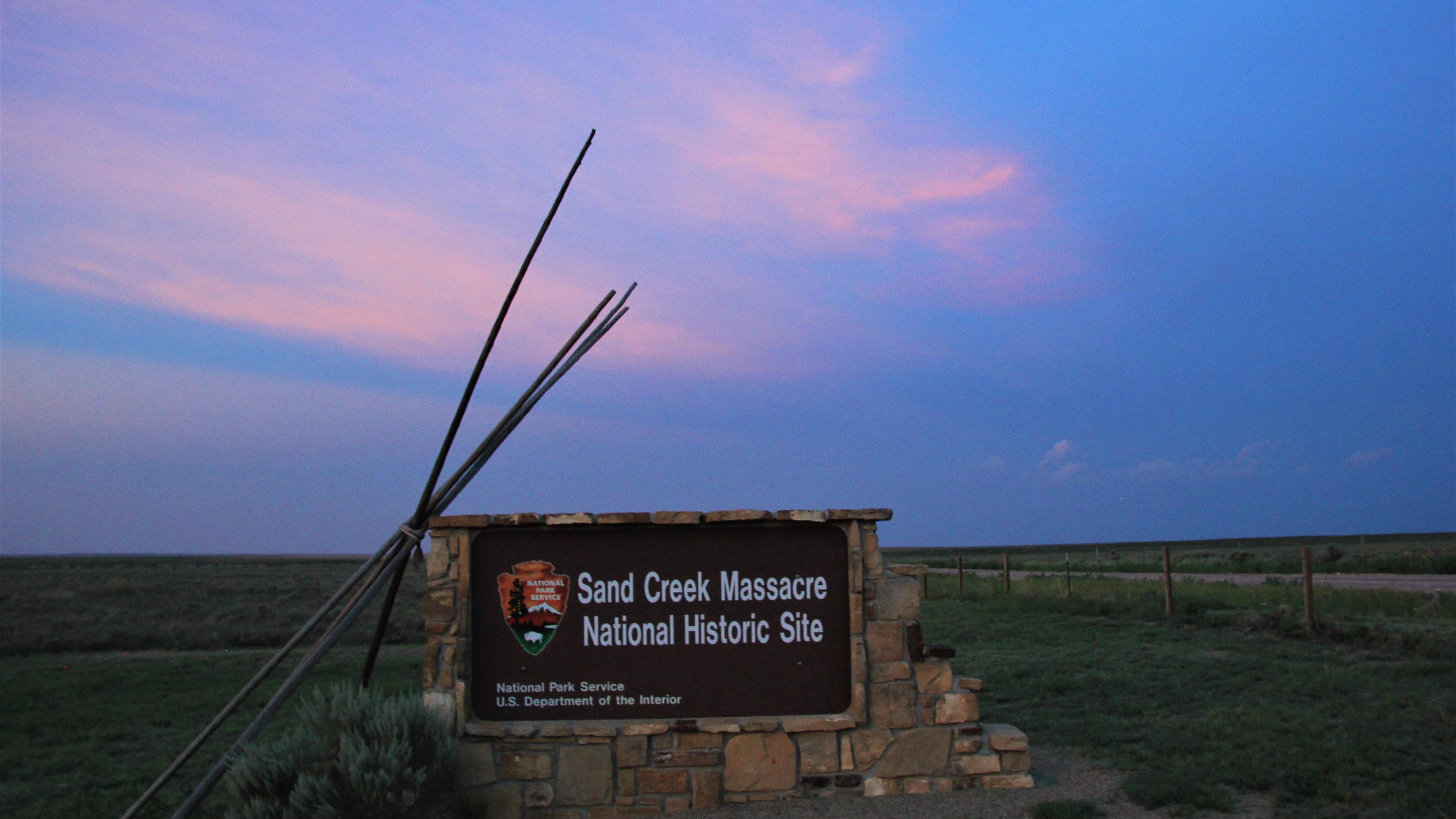 Wood poles lean on a large stone sign in open field at sunset, with dark blue sky and pink clouds.