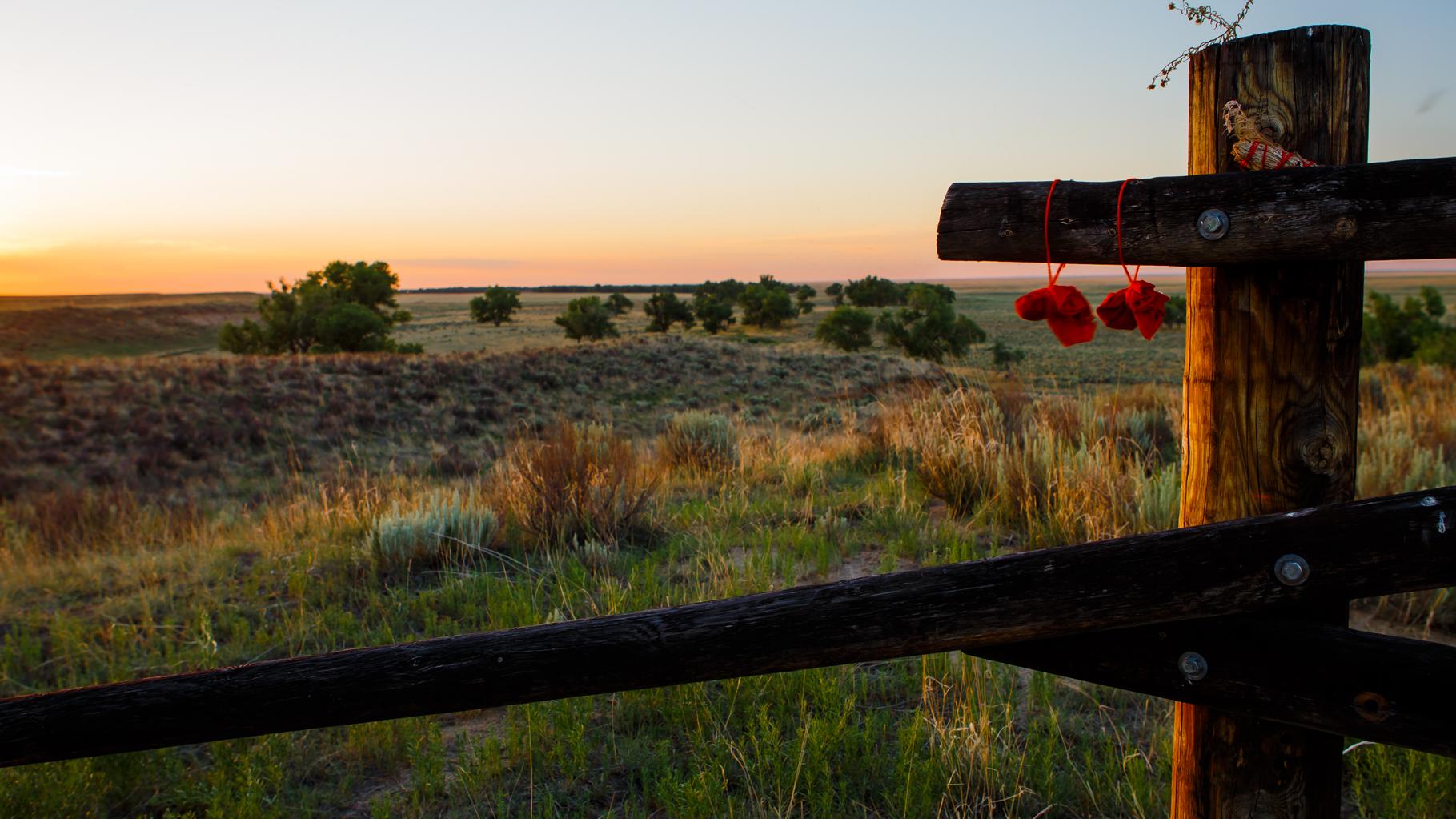 Sand Creek at sunset, framed by a rail fence. In the distance are bluffs and green trees.