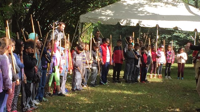 Children standing in line, holding sticks, outside, with grass, trees and a tent visible.  