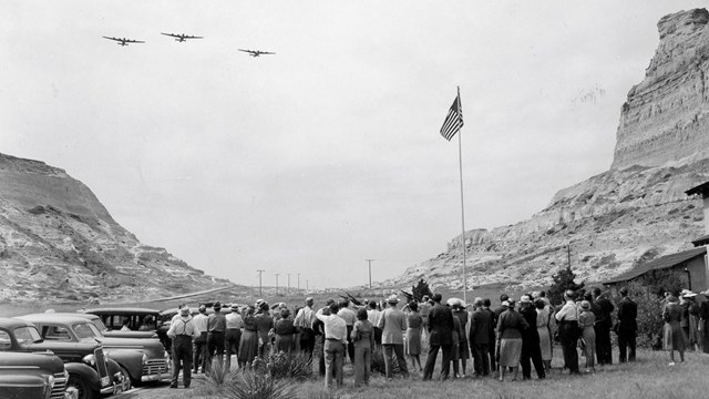 A black and white photo of three airplanes flying over a lawn on which a crowd of people gather.