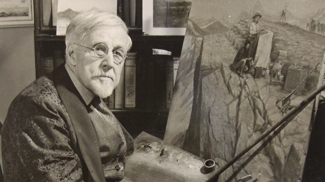 Black and white photo of a man holding a paintbrush and a palette with a painting on an easel.