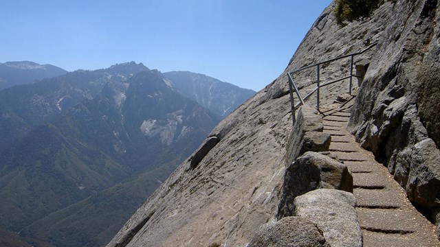 Steps carved into rock and a handrail are pictured right, with mountain viewsheds in left background