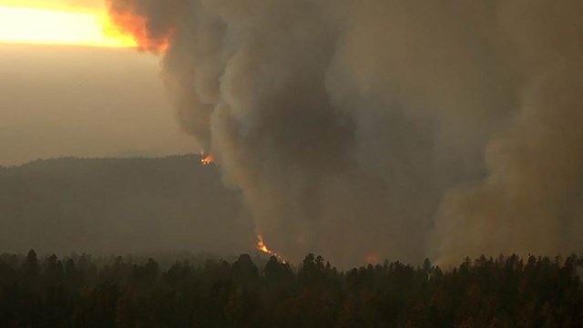 A view of gray smoke rising from a dark forest and a narrow horizontal band of daylight below clouds