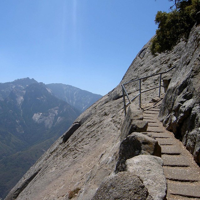 Steps carved into rock and a handrail are pictured right, with mountain viewsheds in left background