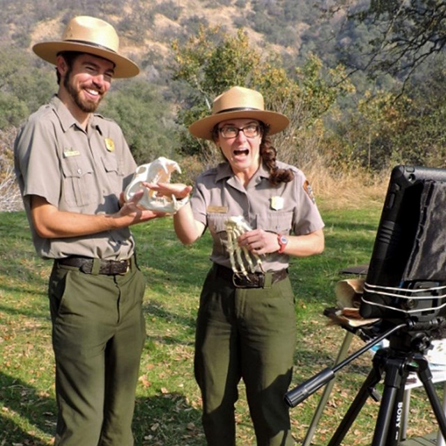 Two rangers stand outside holding a skull. A computer tablet on a tripod faces them.