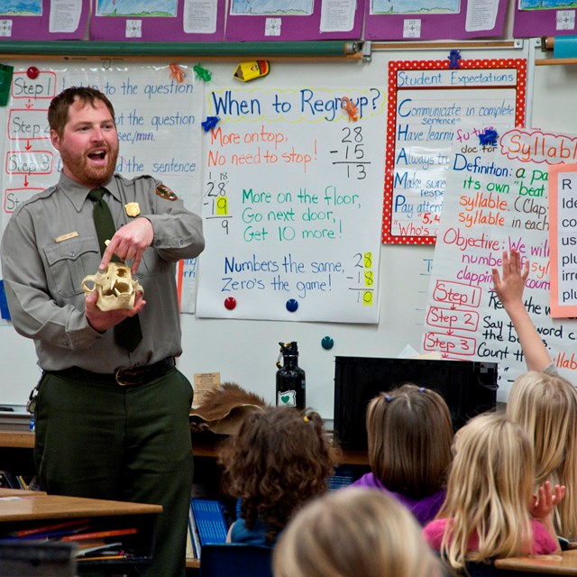 A ranger in the front of a classroom points to a bear skull. One student has raised her hand.