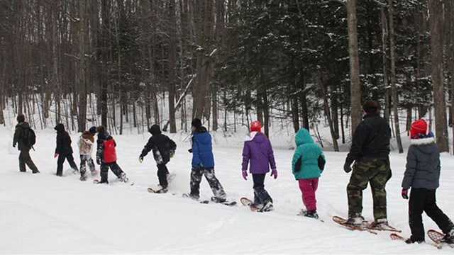 A line of students on snowshoes walk along a snow covered trail