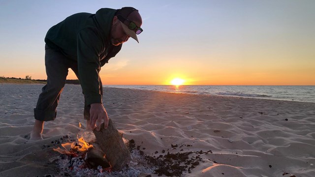 a man leans over a small beach fire to place a log ontop. The sun sets on the horizon over a lake.