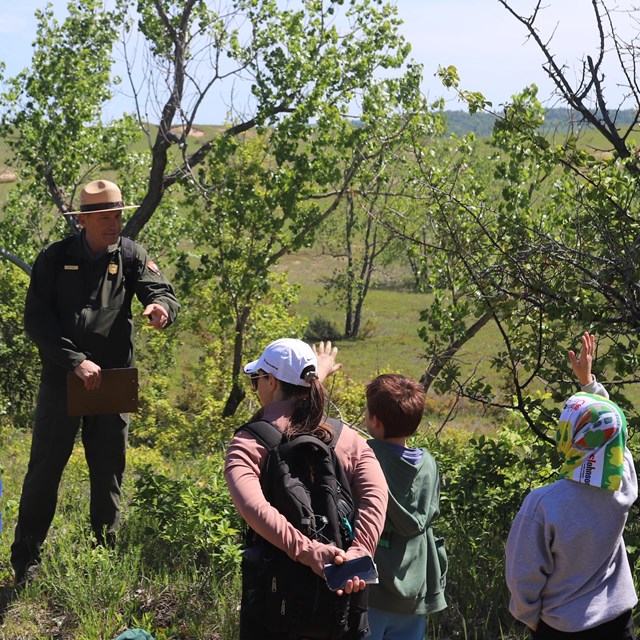 A ranger points to students raising their hands on a field trip. There are trees and grass behind.