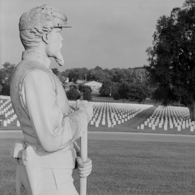 A statue of a soldier stands in a cemetery. A row of graves are in the background.