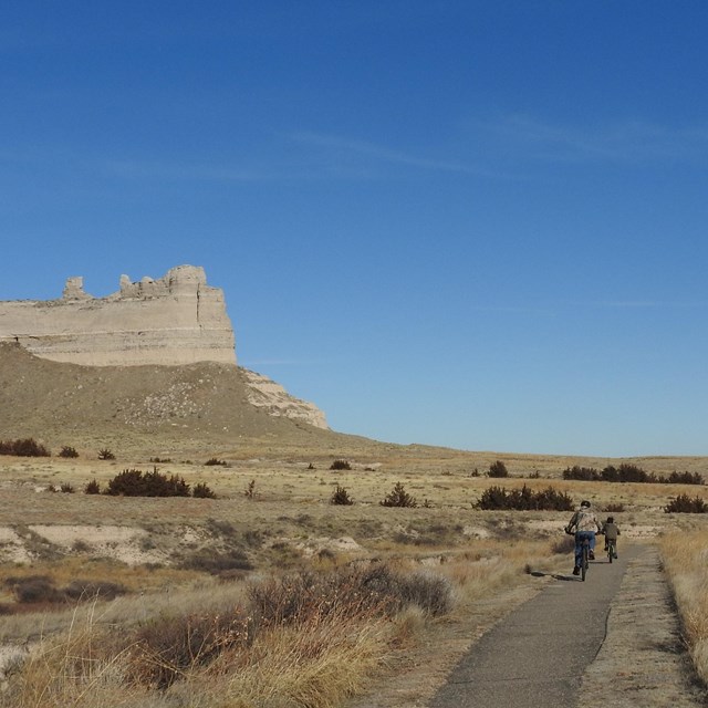 A pair of bicyclists rides on a trail with an impressive rock formation in the background.