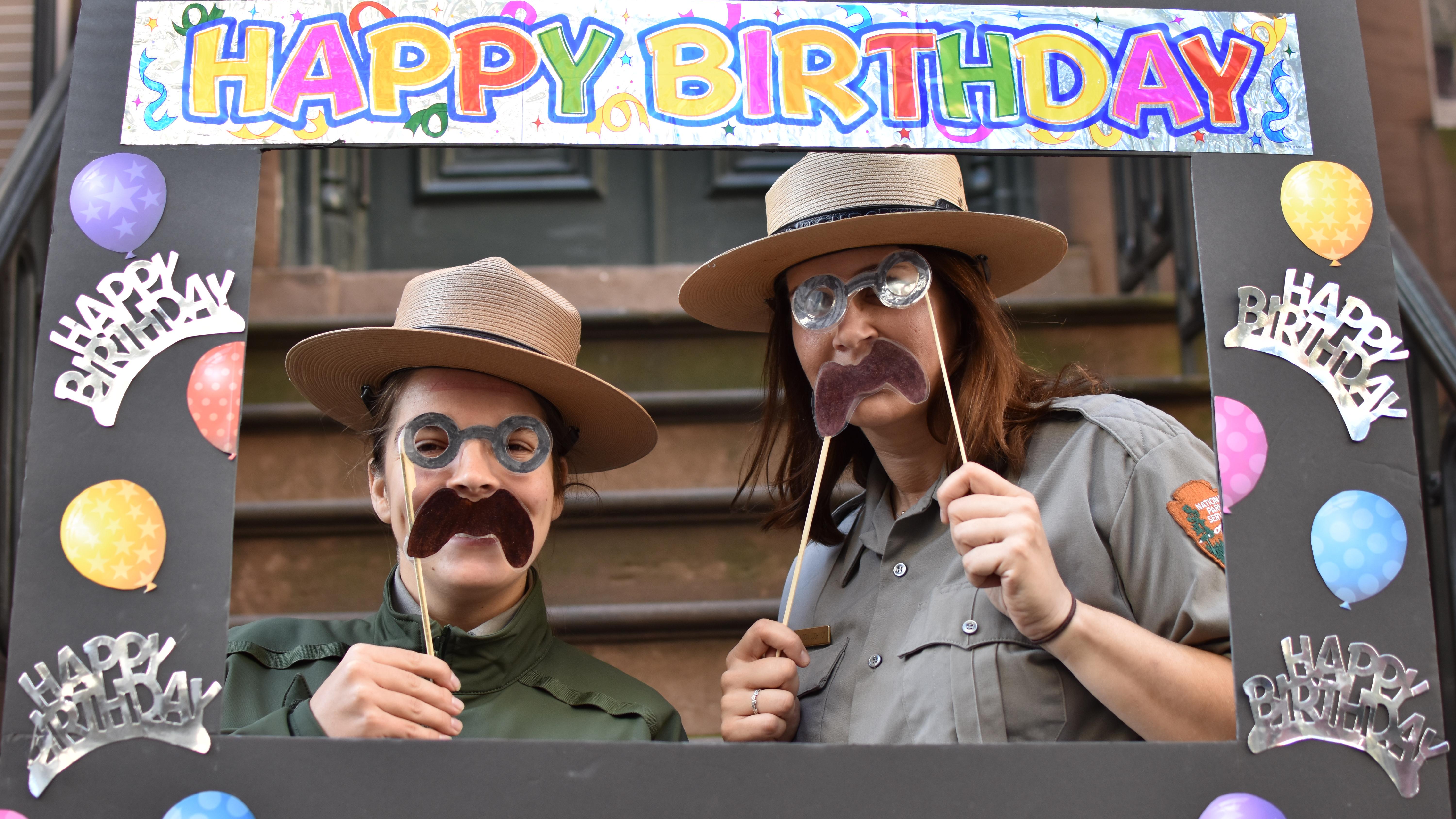 Two rangers holding cut out mustaches and cut out spectacles encircled by a happy birthday frame.