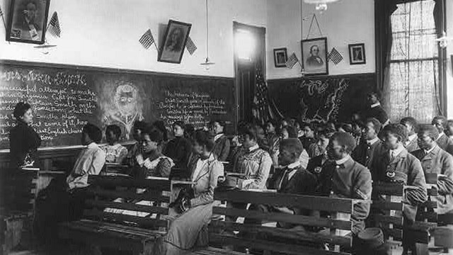  B&W photo of teacher standing in front of classroom of students with writing on blackboard