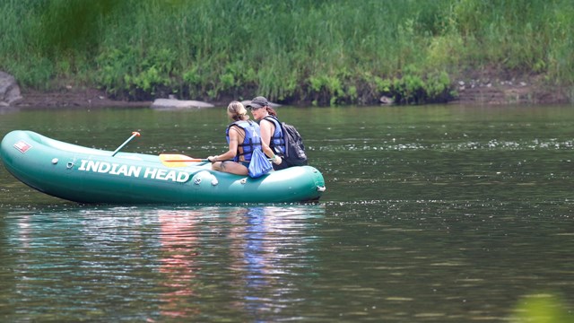 2 people with life jackets in green inflatable raft in river. Raft has printed text "Indian Head."