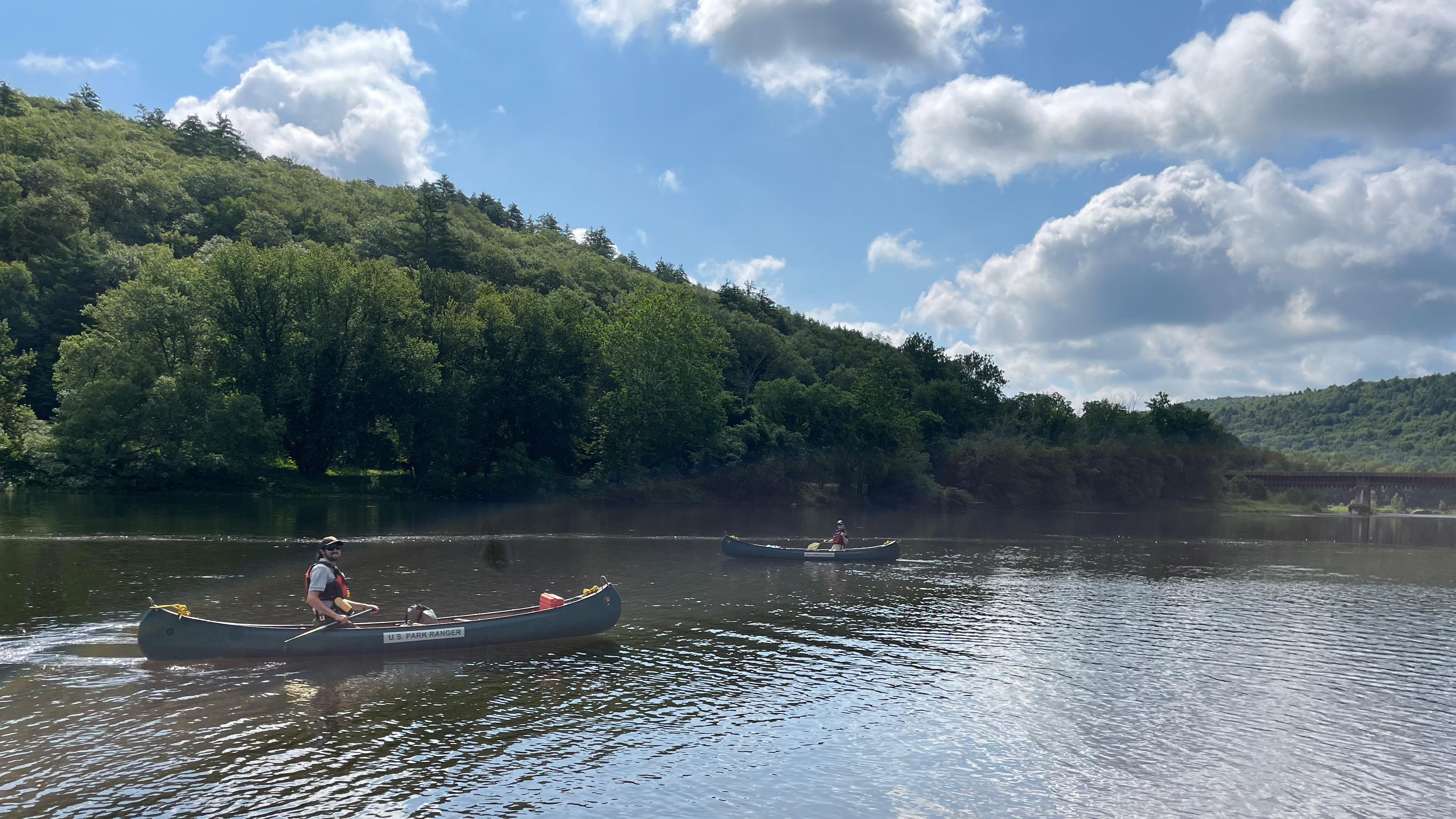 Rangers in canoes on the river. Blue sky with fluffy clouds. Green trees cover hills on river.