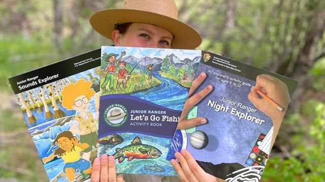 female park ranger holds up three different junior ranger books to the camera
