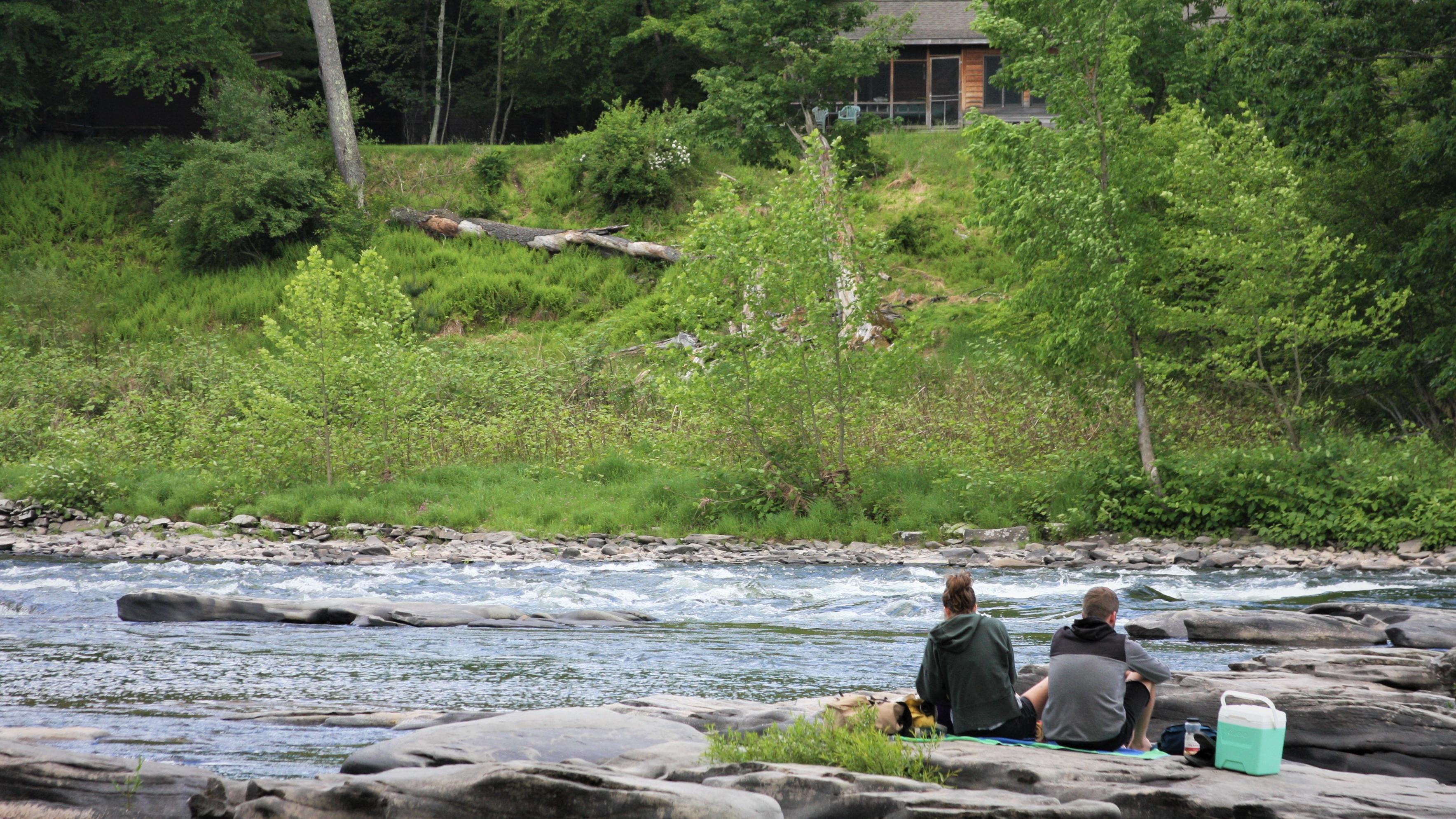 Couple sits on rocks on the edge of a river. Yellow-green trees form a forest across the river.