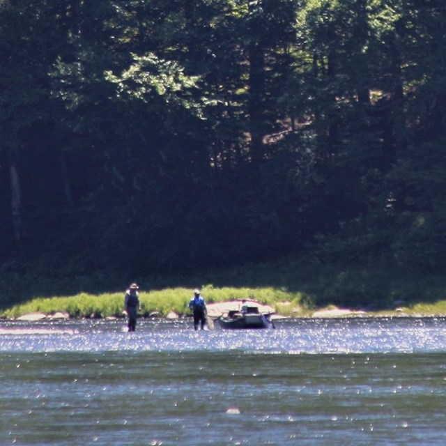 Zoomed out photo of two men standing in shallow part of river next to boat, holding fishing rods. 