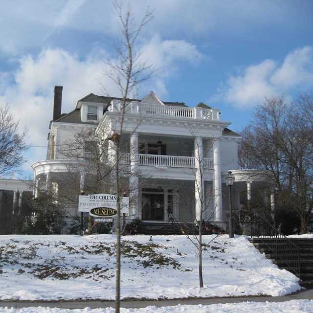 Grand 3-story white building with white columns, large porch and 2nd and 3rd floor railed balconies.