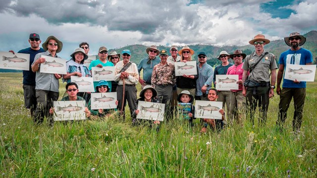 A large group stands in a montane grassland, holding up posters of a native trout.