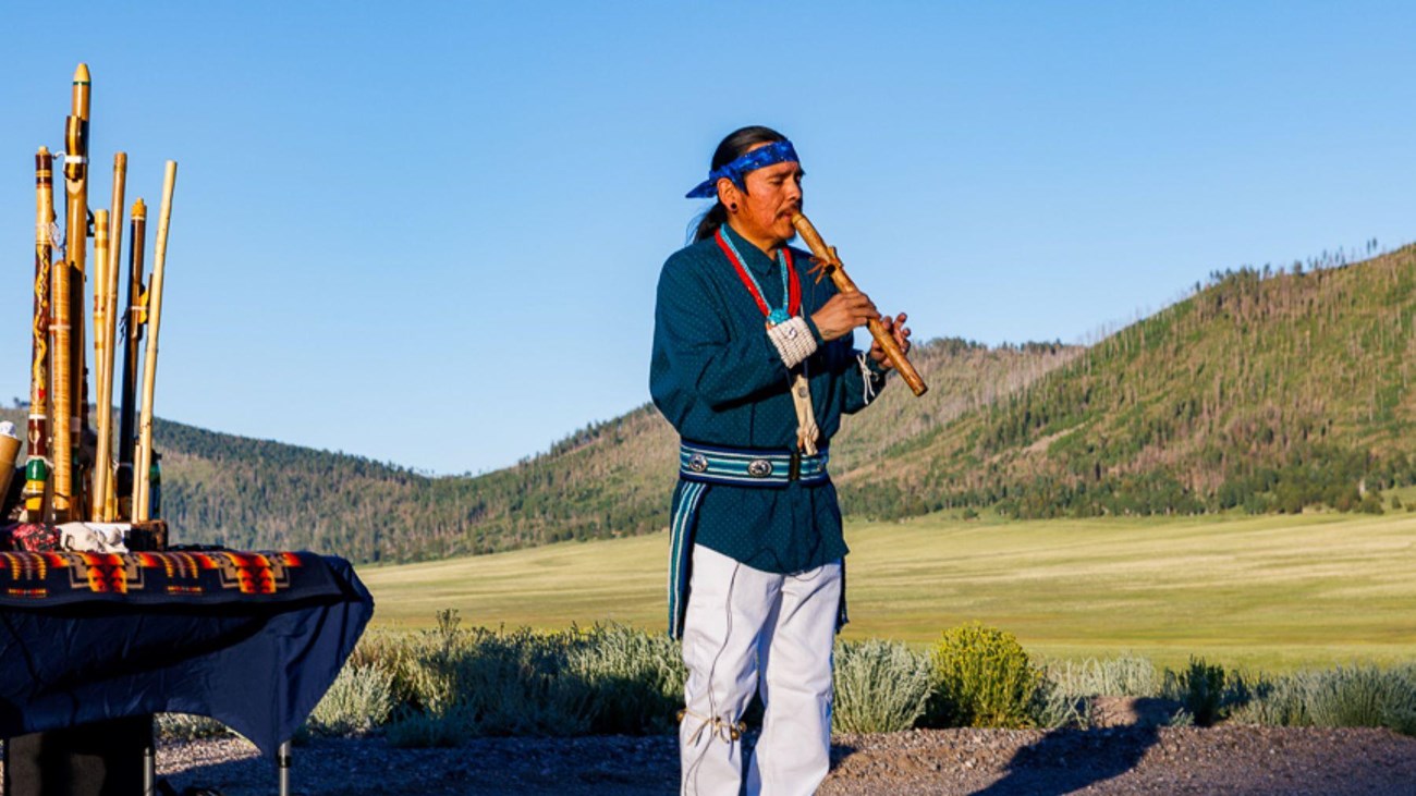 An indigenous man wearing traditional dress plays a wooden flute at the edge of a valley.