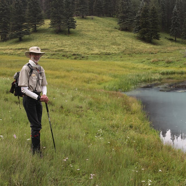 A park volunteer wearing a backpack and a wide-brimmed hat smiles at us from the edge of a pond.