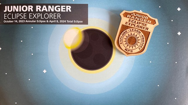Booklet that says Junior Ranger Eclipse Explorer. A wooden badge is on top of the booklet.