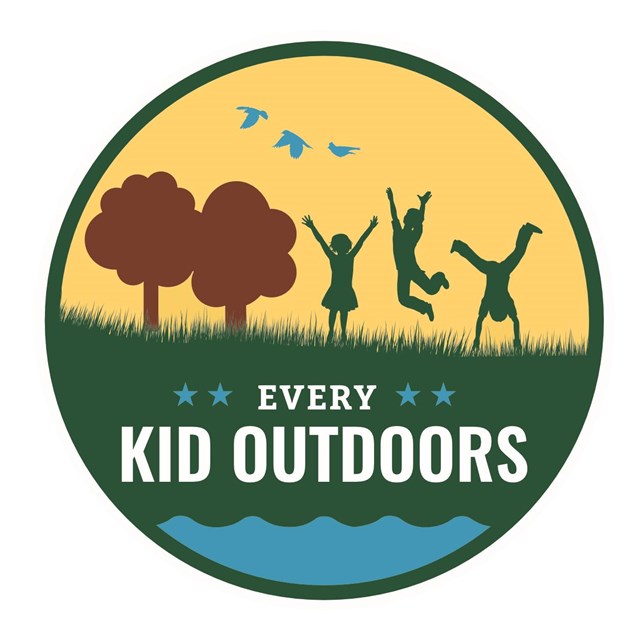 A round logo with kids jumping on green grass, brown trees and yellow sky.