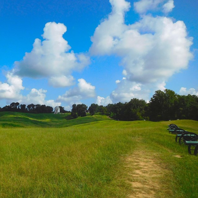 A blue sky with clouds over a green hills with a number of civil war cannons. 