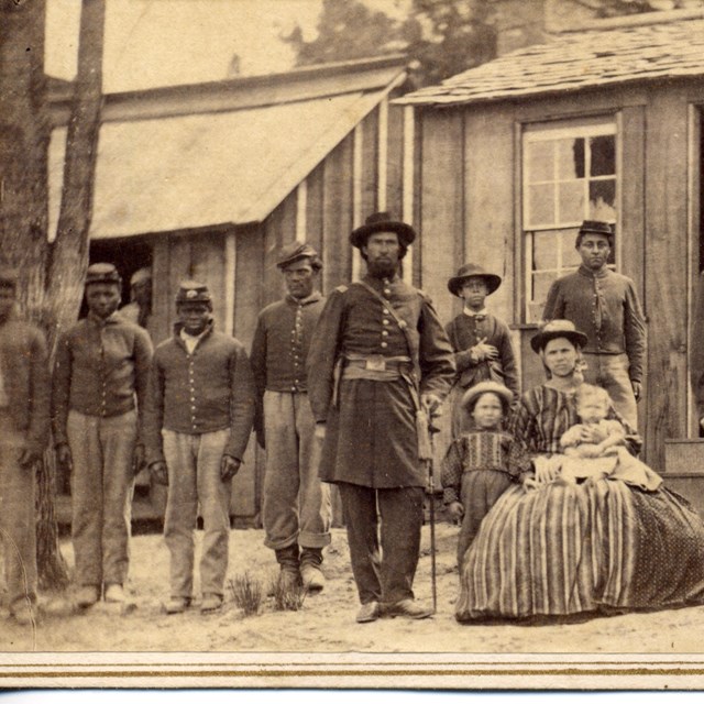 Several African American troops and women in dress stand and sit in front of a building