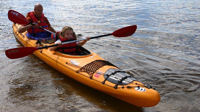 A bright yellow kayak floats near the shore, paddled by a young girl and her father.