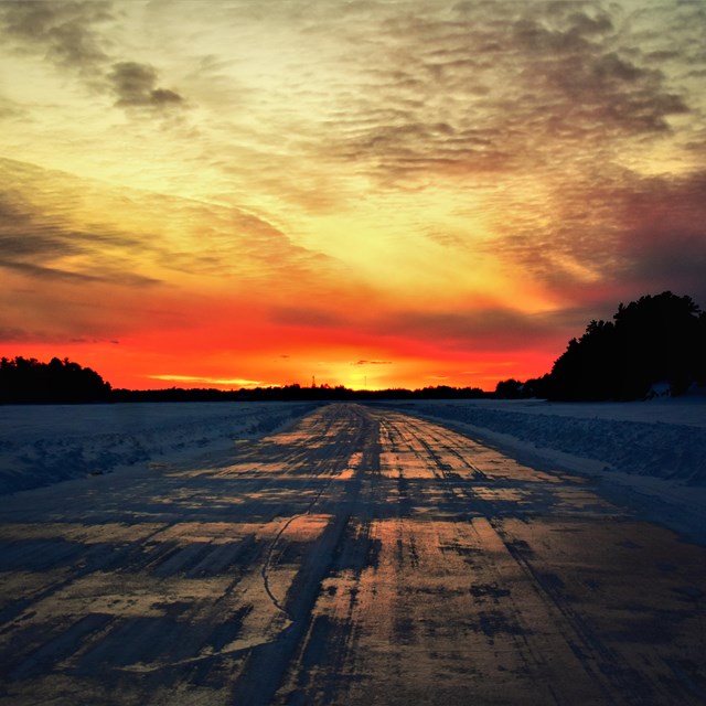 The sun sets bright orange against a dimly lit scenic lakeshore.