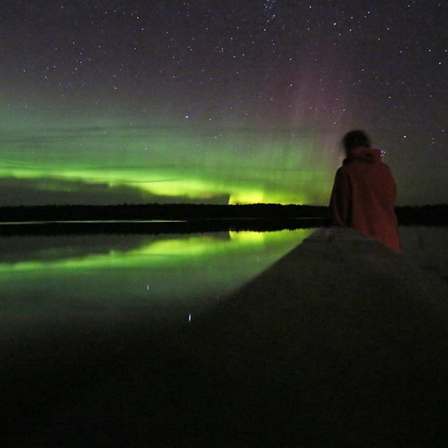 A figure stands at the end of a dock and stares up at a shimmering curtain of green and red lights.