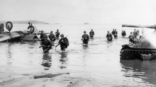 Group of marines wading ashore past airplanes and amtracs