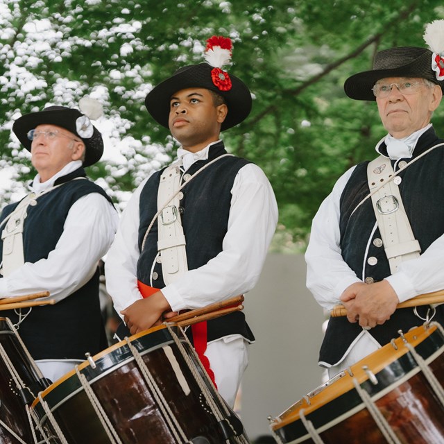 Revolutionary War reenactors in uniform hold drums, standing in formation outdoors.