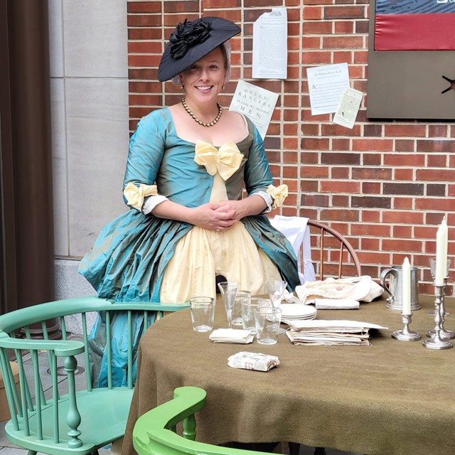 Colonial reenactor in period dress stands by table with candles, papers, and glassware outdoors.