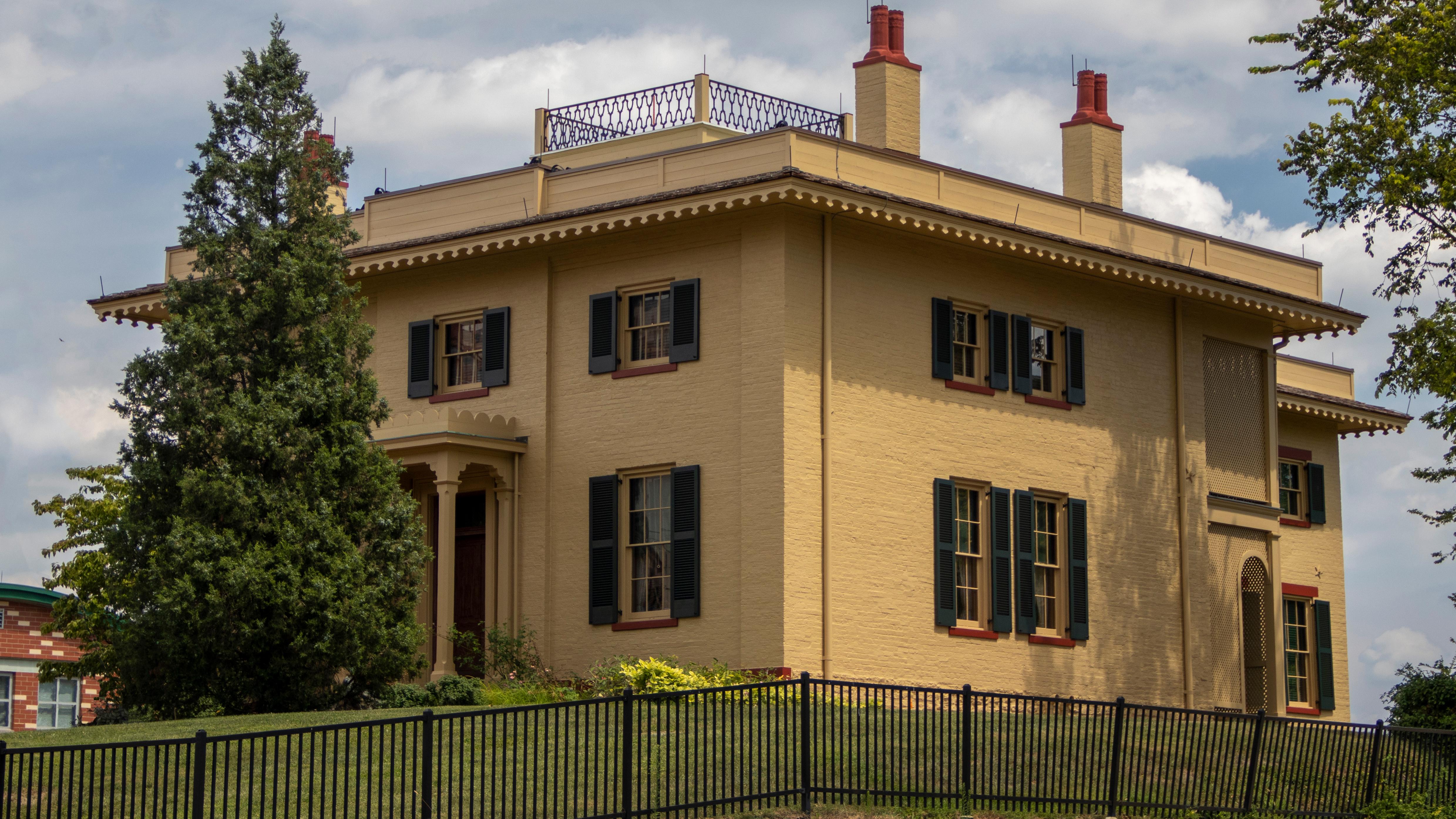 A large, yellow, three-story house with green shutters and several windows with a pine tree in front