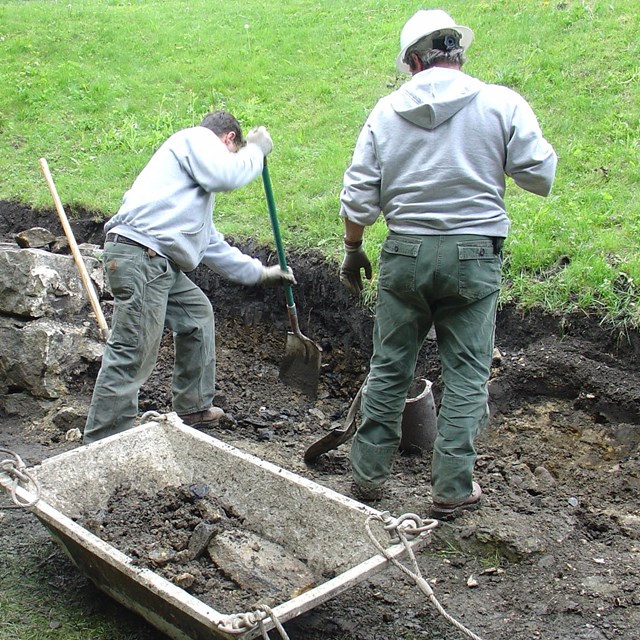 men digging along grassy hill, putting in stone wall