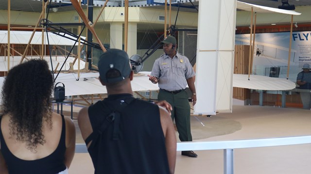 A picture of a male ranger in front of a replica Wright Flyer with two visitors watching