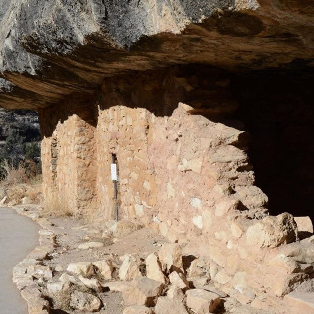 A gravel trail passes in front of a cliff dwelling under a rock overhang