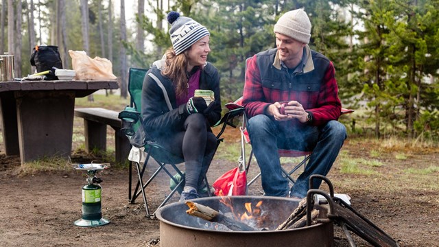 A man and a woman sit by a campfire in a campsite and drink coffee.