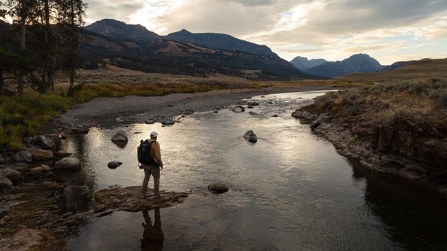 A hiker stands on a rock on the edge of a river in the fading daylight.