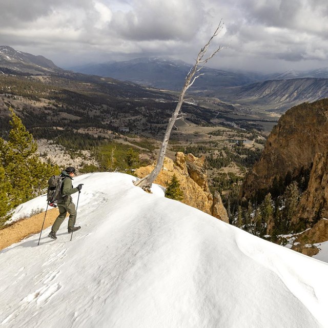 A hiker on an orangish rock outcropping crosses a patch of steep snow.