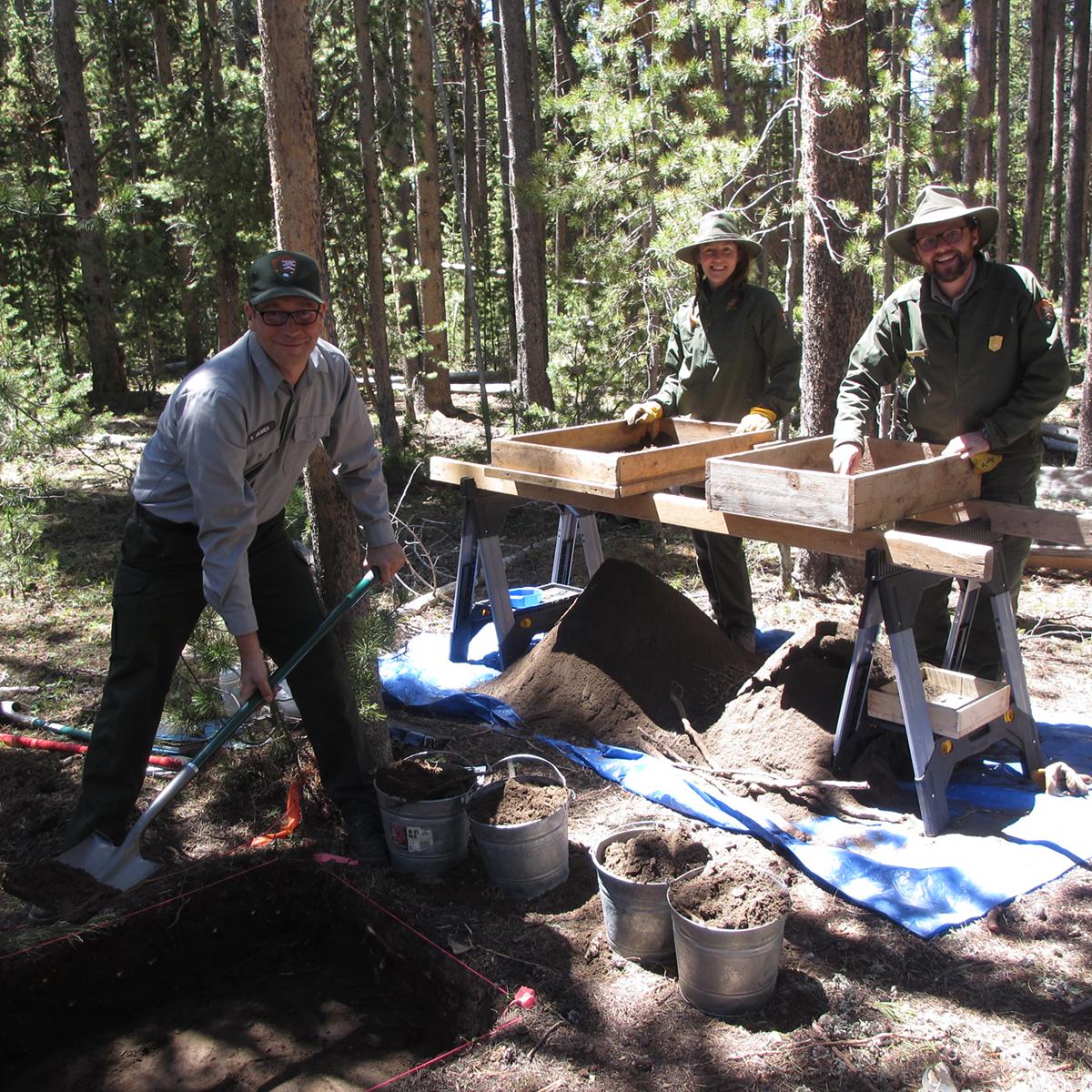 Three uniformed people working around excavated dirt. Two stand by wooden boxes; one uses a shovel.