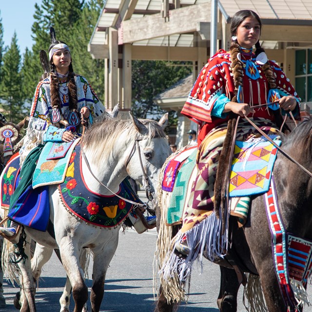 Color photograph showing a line of four horse riders passing in front of the Canyon Village Visitor 